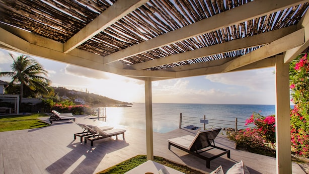 A covered outdoor seating area with lounge chairs and a table, overlooking a body of water. - BEAUTIFUL SUNSET OCEANFRONT VILLA WITH INFINITY POOL (Crosbies)