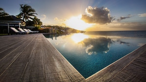 A wooden infinity pool with clear water reflecting the sky and sun, lounge chairs, and a palm tree. - BEAUTIFUL SUNSET OCEANFRONT VILLA WITH INFINITY POOL (Crosbies)