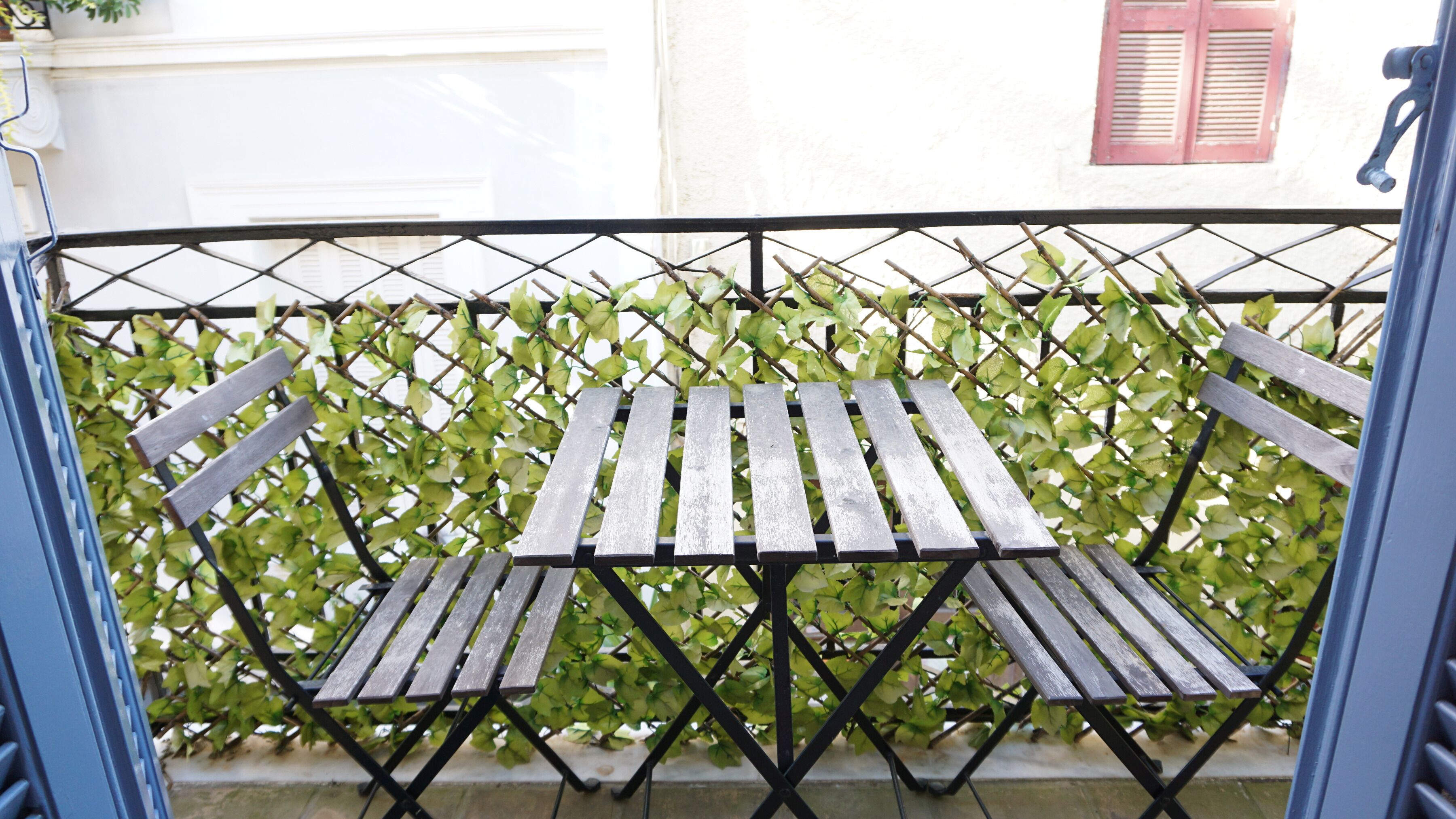 Ein Tisch mit Stühlen steht auf einem Balkon, der von einer Gitterzäunung und grünem Laubwerk gesäumt wird.
