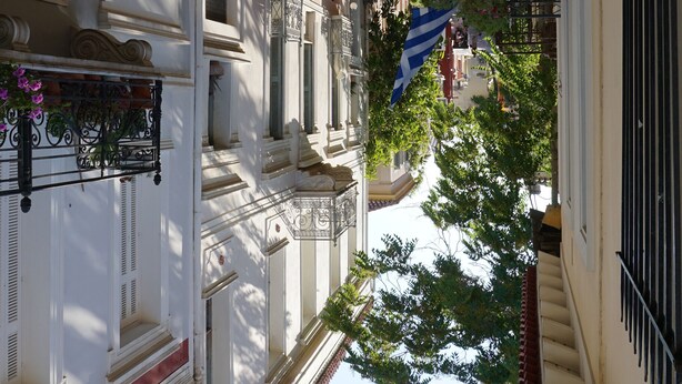 Una calle estrecha flanqueada por edificios, con balcones adornados con plantas en macetas y una bandera azul.