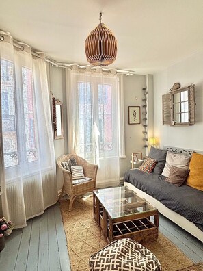 Un salón acogedor con un sofá, un sillón de mimbre, una mesa de centro de vidrio y un reposapiés estampado.