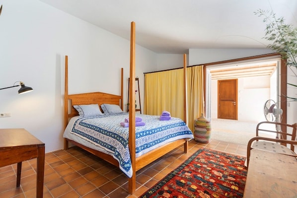 Ein Zimmer mit einem Himmelbett, einem Holztisch, einem Stuhl, einem Ventilator und einer großen Vase.