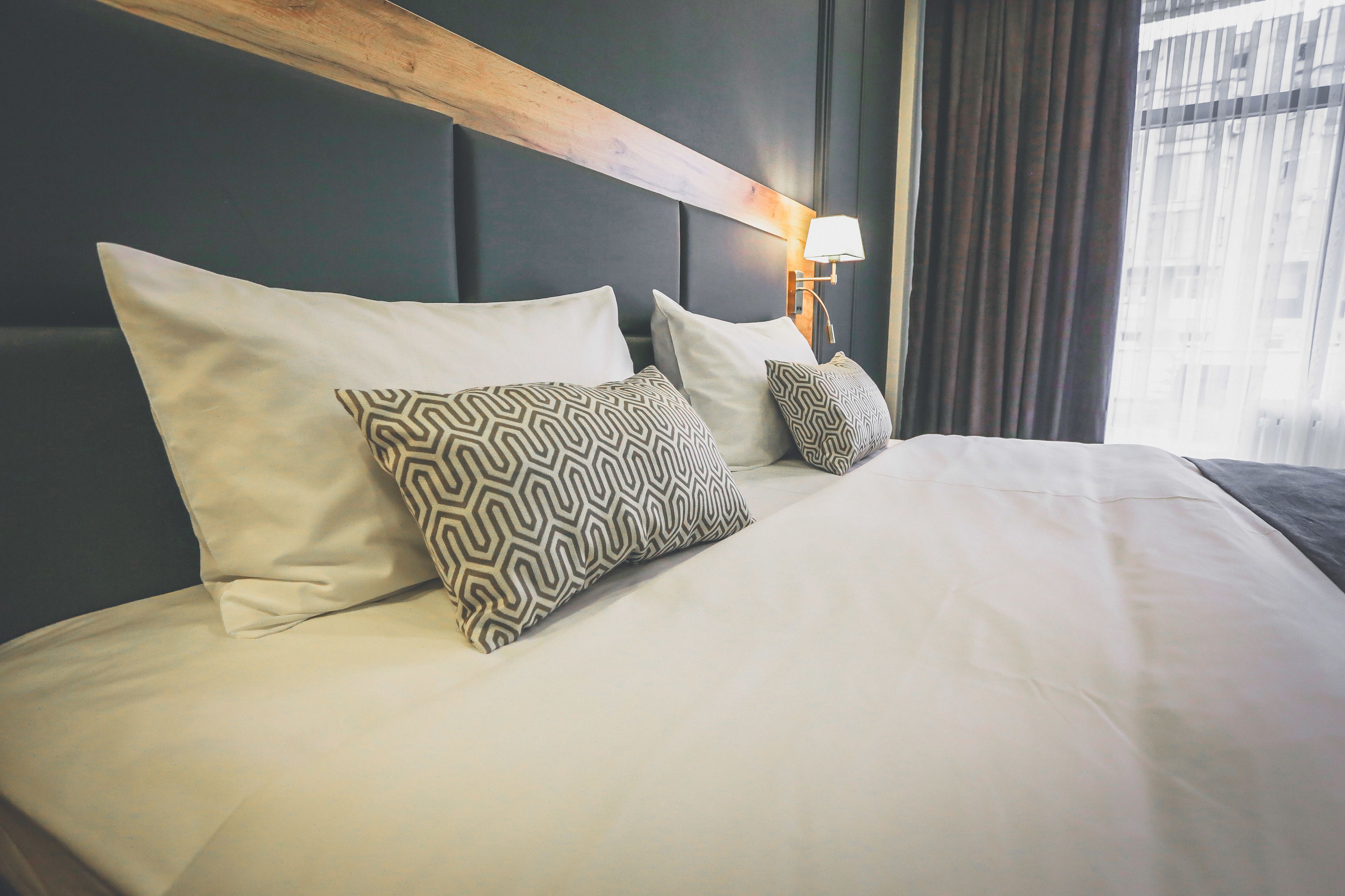 Una cama bien hecha con almohadas estampadas, un cabecero de madera y una lámpara de pared.