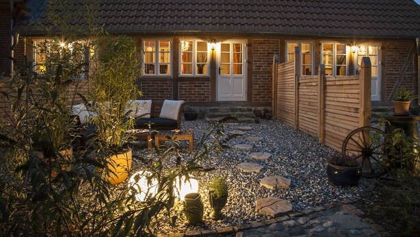 A cozy cottage with a tiled roof, a wooden fence, and a gravel patio with outdoor furniture.