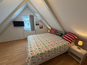Un dormitorio con techo inclinado, una cama con ropa de cama estampada, una mesita de noche con lámpara y una ventana con cortinas.