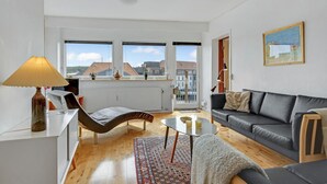 Ein modernes Wohnzimmer mit einer Couch, einem Sessel, einem Tisch und einer Lampe. Drei Fenster bieten Blick auf Gebäude, ein Heizkörper ist ebenfalls vorhanden.