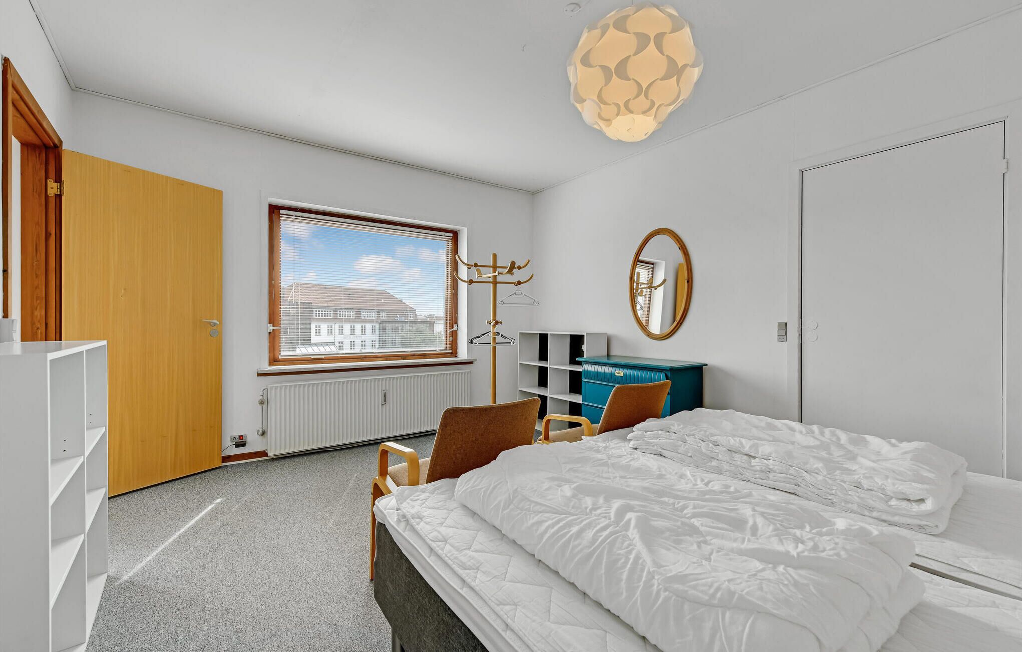 A modern bedroom with a large bed, a desk, a chair, a coat rack, and a view of buildings through the window.