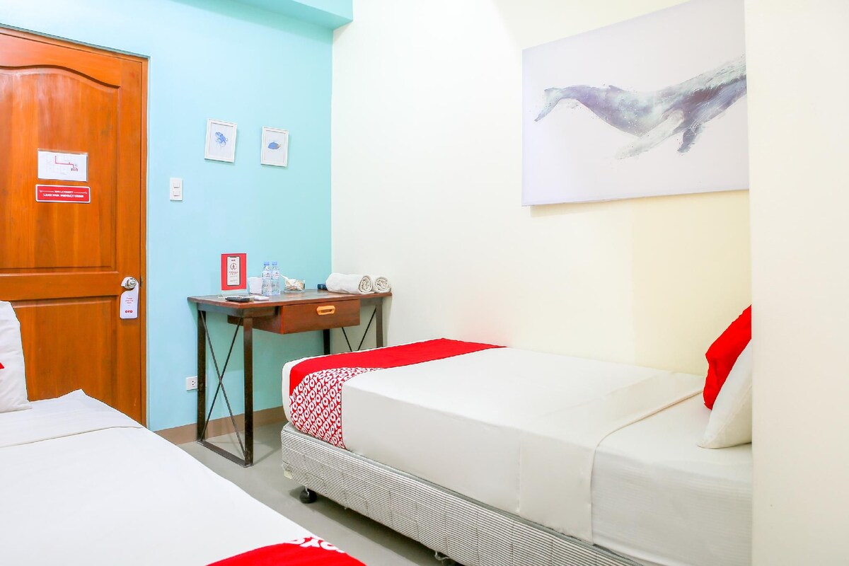 A hotel room with a bed, a desk with a chair, a small table, a wall-mounted artwork, and a door with a sign.