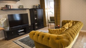 Ett vardagsrum med en platt-TV, en svart möbel för underhållningsutrustning, en soffa i senapsgul färg och ett fönster med gardiner.
