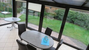 A patio area with a round table and four chairs, large windows offering a view of a garden, and a ceiling with a textured pattern. - Vacation home Gala in the vacation village Altes Land - Holiday home 303 Gala 80sqm for max. 6 persons (Hollern-Twielenfleth)