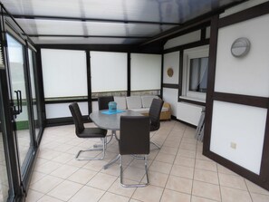 A covered patio with a round table and six chairs, a sofa, and a window with blinds.