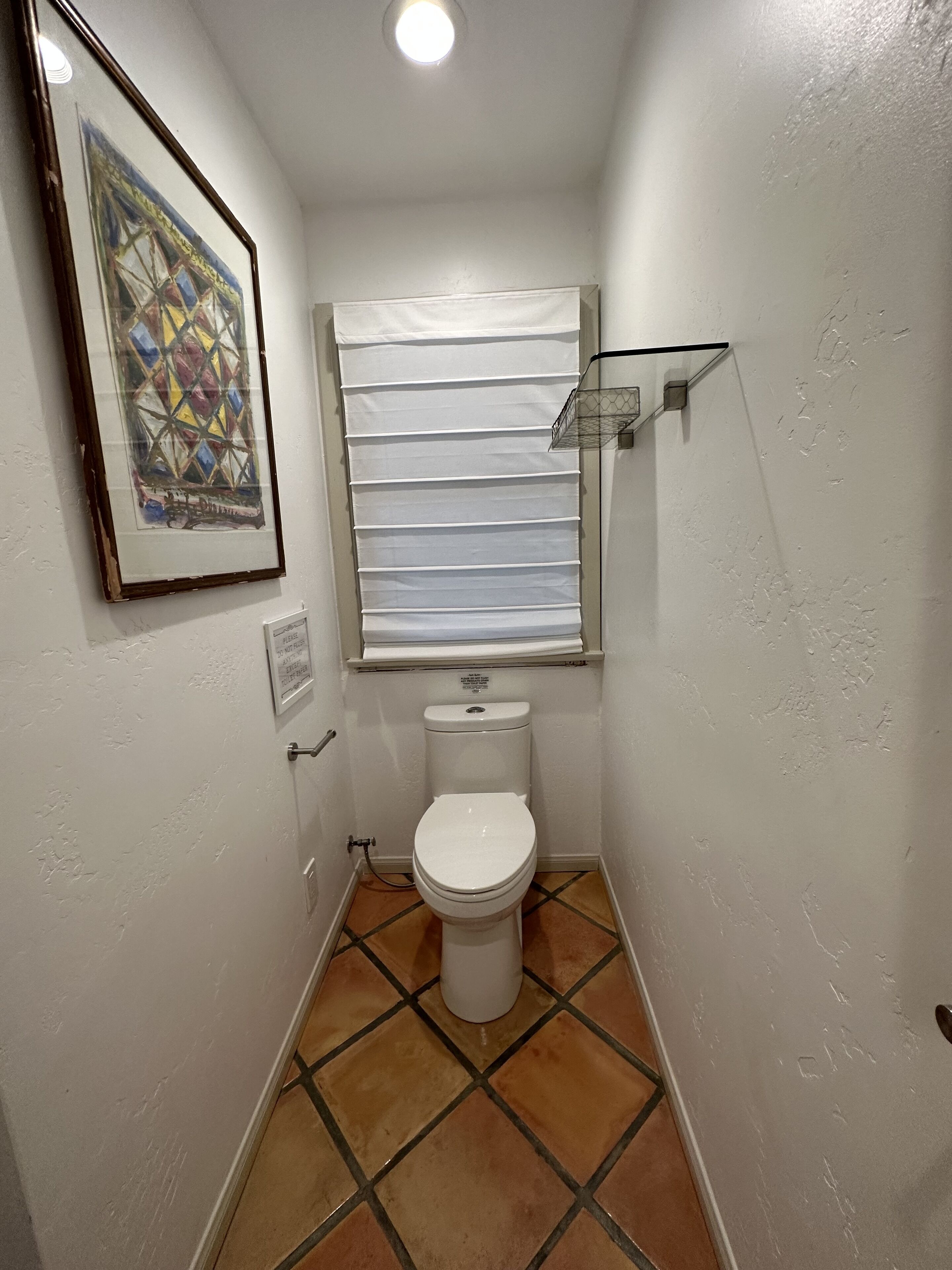 A small bathroom with a toilet, a window with blinds, a framed picture on the wall, and a tiled floor.