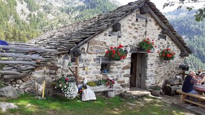 A rustic stone building with a sloped roof, wooden benches, and flower baskets. - Lovely little cottage in the alpine pastures (Lillianes, Vallée d'Aoste)