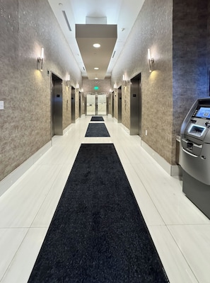 Corredor de hotel com carpete preto, elevadores e um quiosque de autoatendimento Samsung.