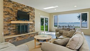 Ein Wohnzimmer mit Kamin aus Stein, einem Flachbildfernseher, einem großen Fenster mit Blick auf das Wasser und einem beigefarbenen Ecksofa.