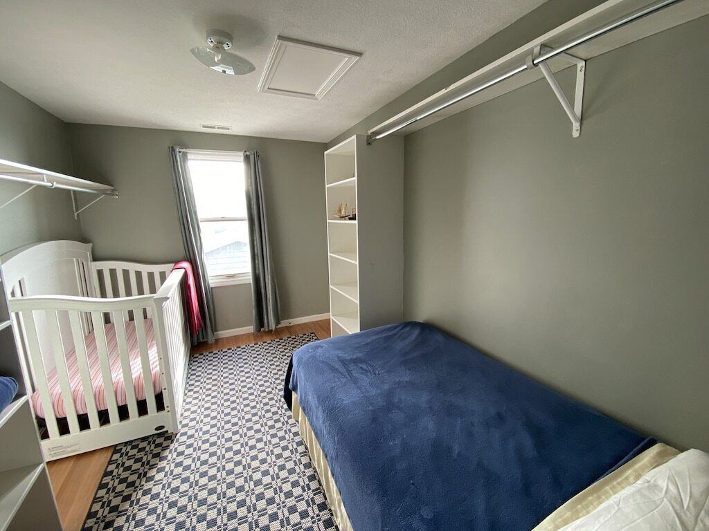A room with a crib, a bed, a checkered floor, a window with curtains, and a shelf unit.