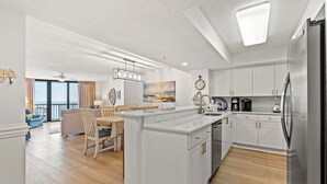 A modern kitchen with white cabinetry, a central island, and a dining area with a view. - Naturally Beautiful Unit, Recliner, Indoor Pool | Ocean Bay Club - 807 (North Myrtle Beach)