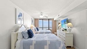 A bedroom with two beds, a ceiling fan, a dresser with lamps, and a large window with a view. - Naturally Beautiful Unit, Recliner, Indoor Pool | Ocean Bay Club - 807 (North Myrtle Beach)
