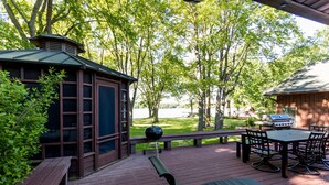 Una terraza de madera con una parrilla, una mesa de picnic y sillas, rodeada de árboles.