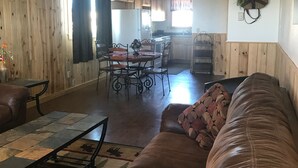 Un salon chaleureux avec un canapé en cuir marron, un coin repas avec une table et des chaises, des murs en bois et une cuisine à l’arrière-plan.