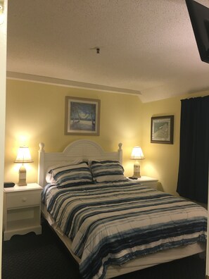 Une chambre à coucher avec un lit, deux tables de chevet avec des lampes, un luminaire au plafond et deux tableaux encadrés accrochés au mur.