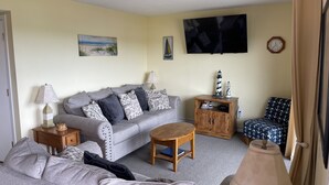 A living room with a grey sofa, a wooden coffee table, a TV mounted on the wall, a small cabinet with decorative items, and a patterned armchair. - Myrtle Beach Resort, Ocean View, 2 BR - 2 BA, Indoor/Outdoor Pool, B-244 (Myrtle Beach)