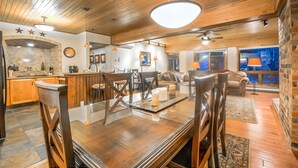 A spacious dining area with a wooden table and chairs, a kitchen in the background, and a large window with a view.