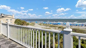 A beach view from a balcony with a white railing, palm trees, and a sandy beach with blue umbrellas.