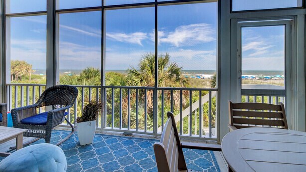 A covered outdoor area with a view of the beach, furnished with a chair, table, and a potted plant.