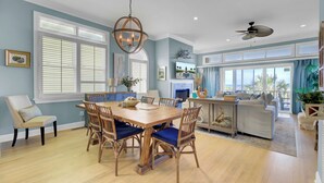 A spacious dining area with a wooden table and chairs, a chandelier, and large windows with blinds.