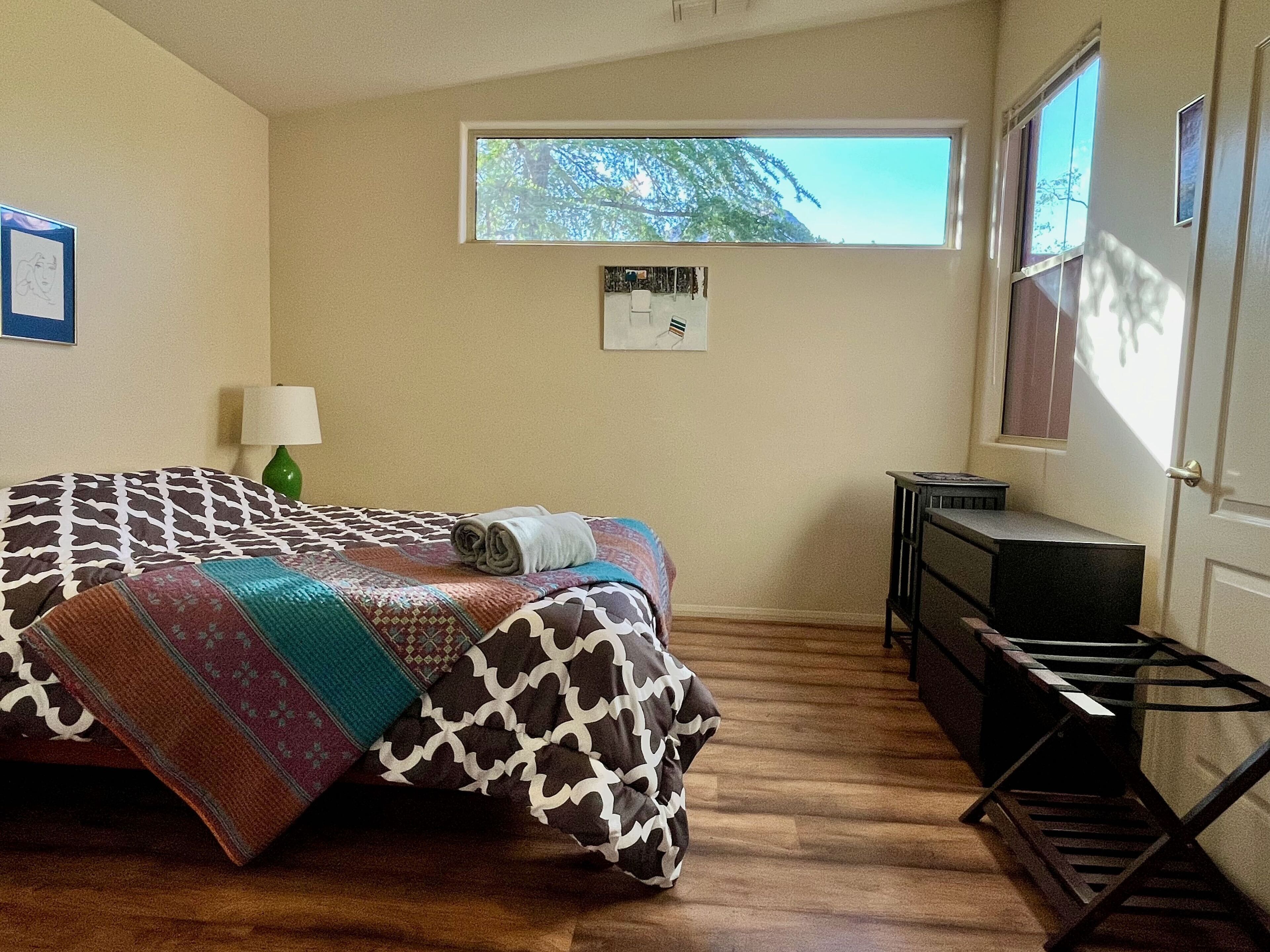 A bedroom with a bed, a desk, a lamp, and a window with a view of trees.