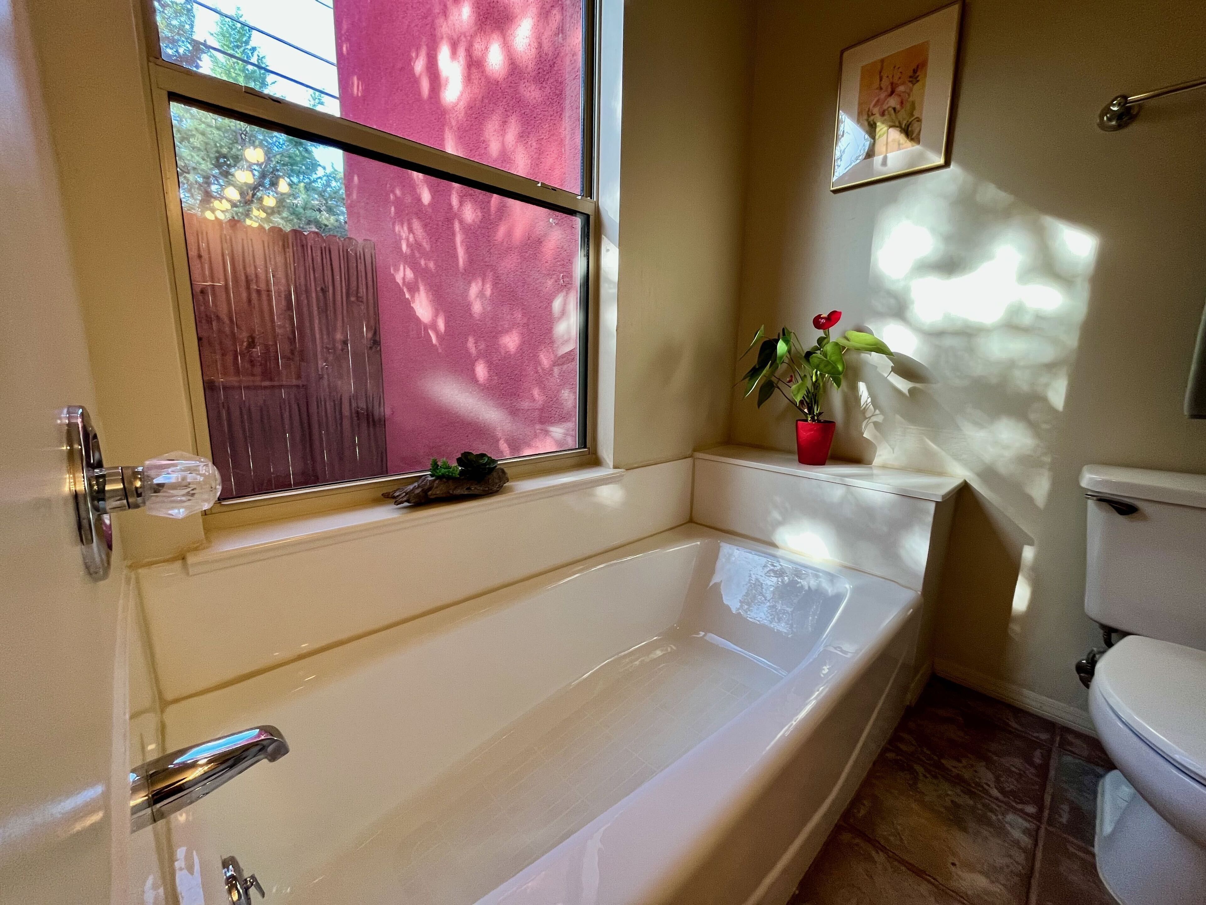 A bathroom with a bathtub, a toilet, and a window with a view of a wooden fence and trees.