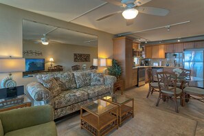 A living room with a sofa, coffee table, and chairs, a dining area with a table and chairs, and a kitchen with wooden cabinets and appliances. - Leinaala Oceanfront 405 Panoramic Ocean View, Great Rates, Steps to the beach (Kihei)