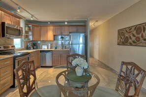 A modern kitchen with wooden cabinets, stainless steel appliances, and a dining area with a glass table and chairs. - Leinaala Oceanfront 405 Panoramic Ocean View, Great Rates, Steps to the beach (Kihei)