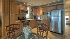 A modern kitchen with wooden cabinets, stainless steel appliances, and a dining area with a glass table and chairs. - Leinaala Oceanfront 405 Panoramic Ocean View, Great Rates, Steps to the beach (Kihei)