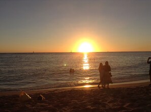 A beach scene at sunset with the sun low on the horizon, people walking along the shore, and a sailboat in the distance.