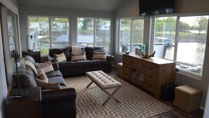 A spacious living room with a dark leather sofa, a wooden cabinet, and a large window providing a view of the outdoors. - GRAND PANORAMIC RETREAT, 6 BR, 3 BA, 300' Lakefront, Dock & Launch! NEW (Grove)
