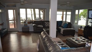 A spacious living room with a leather sofa, a coffee table, and a television. There are large windows providing a view of the outdoors. - GRAND PANORAMIC RETREAT, 6 BR, 3 BA, 300' Lakefront, Dock & Launch! NEW (Grove)
