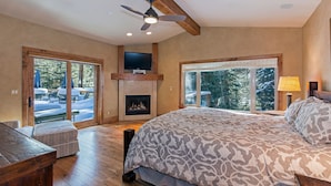 Un dormitorio con una cama grande, una chimenea, un televisor montado encima y vistas del exterior a través de amplios ventanales.