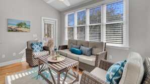 A sunlit room with a ceiling fan, a sofa with cushions, a wicker chair, a wooden table with a tray, and a window with blinds. - Award-Winning Resort Amenities | Back Bay 5813 (Isle Of Palms)
