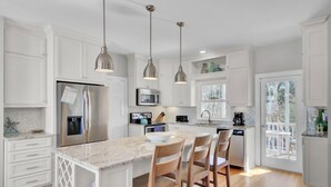 A modern kitchen with white cabinetry, a central island with bar stools, stainless steel appliances, pendant lighting, and a view to a deck. - Award-Winning Resort Amenities | Back Bay 5813 (Isle Of Palms)