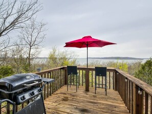A wooden deck with a barbecue grill and two chairs under a red umbrella, overlooking a body of water. - Romantic Lakeview Treehouse with Private Jacuzzi (Stigler)