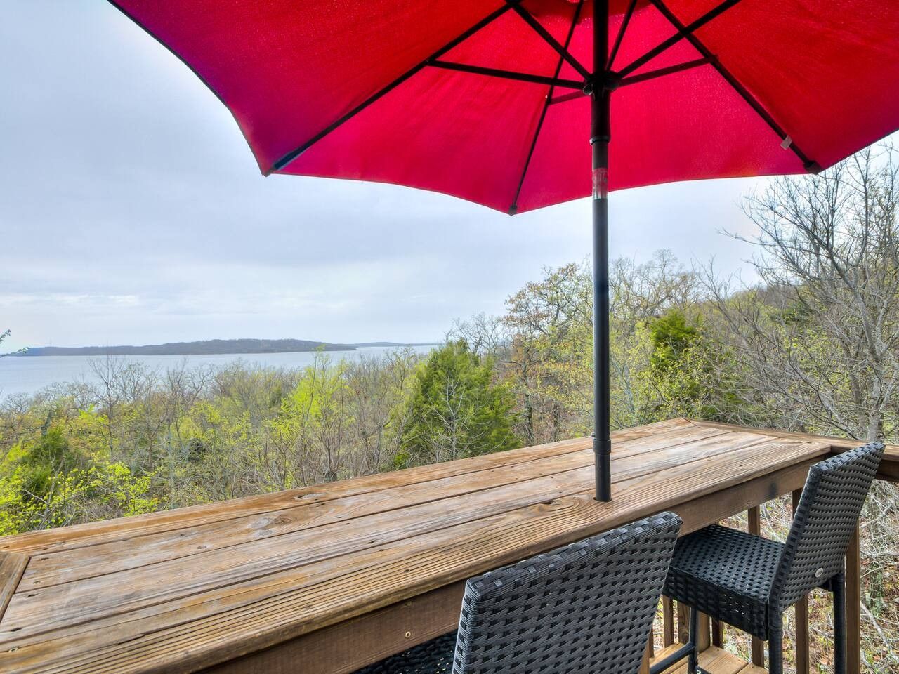 A wooden deck with a red umbrella, a table, and two wicker chairs overlooking a body of water.