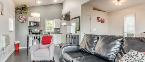 Un salon moderne avec un canapé en cuir, un fauteuil blanc et un coin repas, la cuisine étant visible au fond.