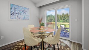 A dining area with a glass table, four chairs, a floral rug, a painting, and a view of a balcony with outdoor furniture. - Stylish Two-Story Getaway on Keizer's Quiet Outskirts (Keizer)