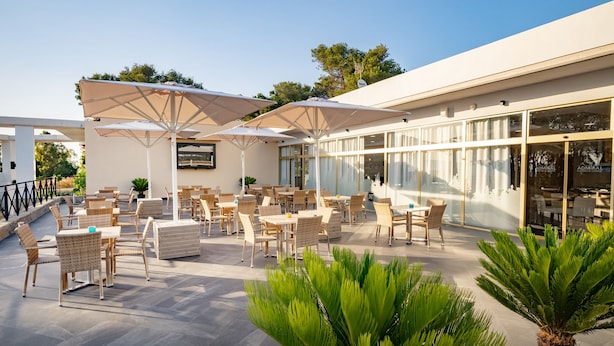 Een terras met tafels en stoelen, afgedekt door parasols, en een gebouw met grote ramen op de achtergrond.