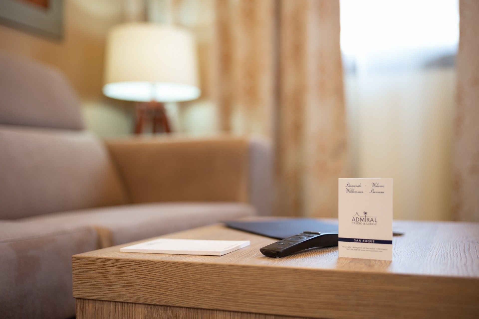 A hotel room with a wooden nightstand, a remote control, and a welcome card.