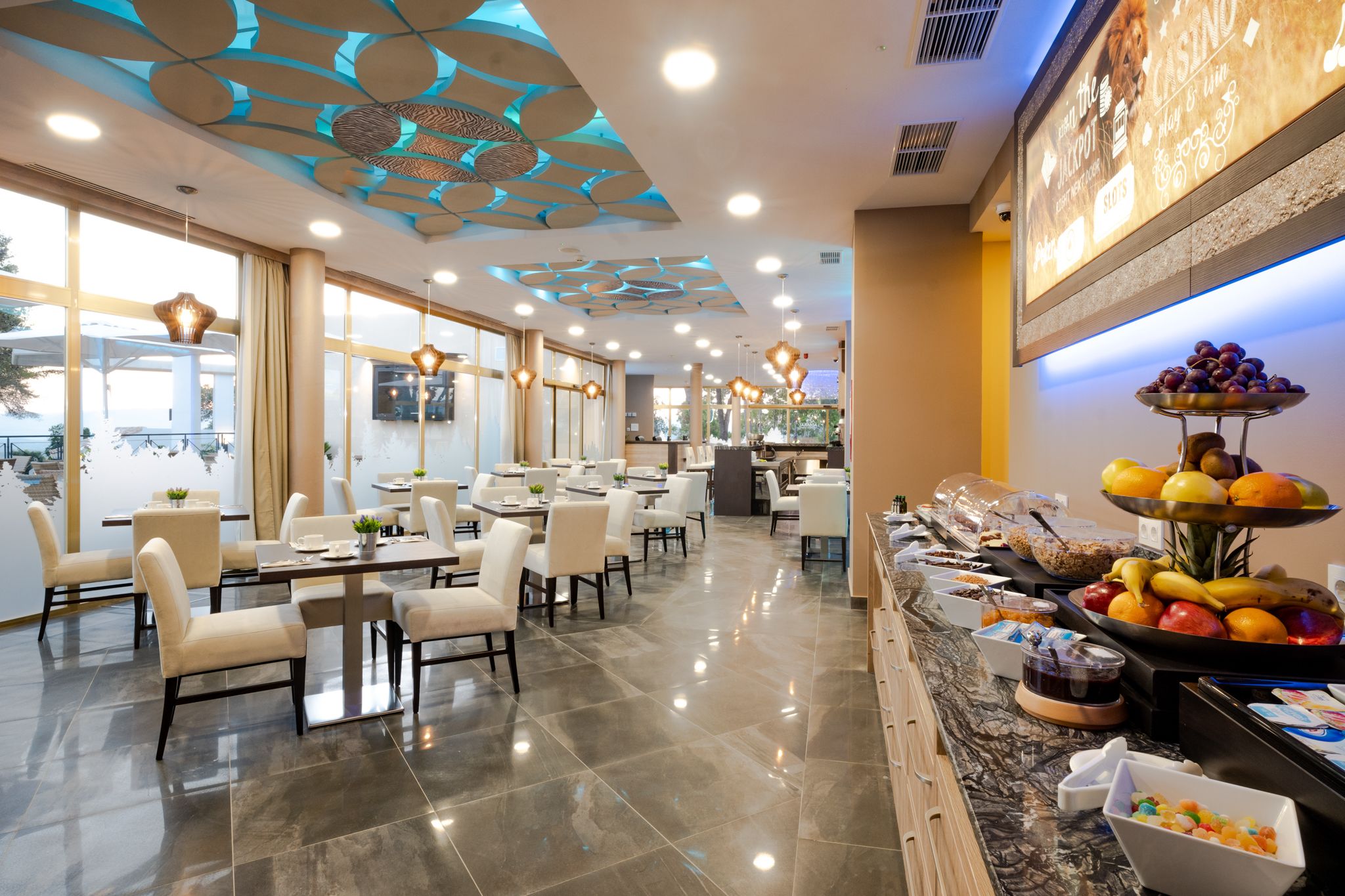 A modern hotel dining area with a buffet, large windows, and a decorative ceiling.