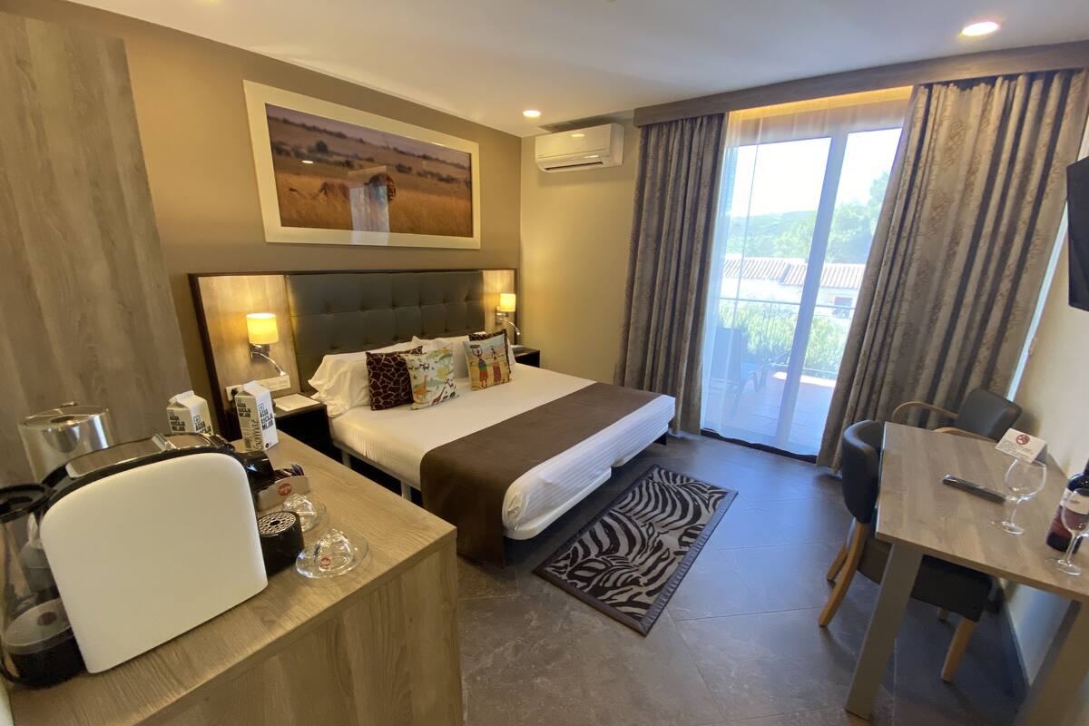 Une chambre d’hôtel avec un lit, des tables de chevet, un bureau, une chaise, un tapis à motifs zébrés, un tableau au mur et une vue à travers les rideaux.