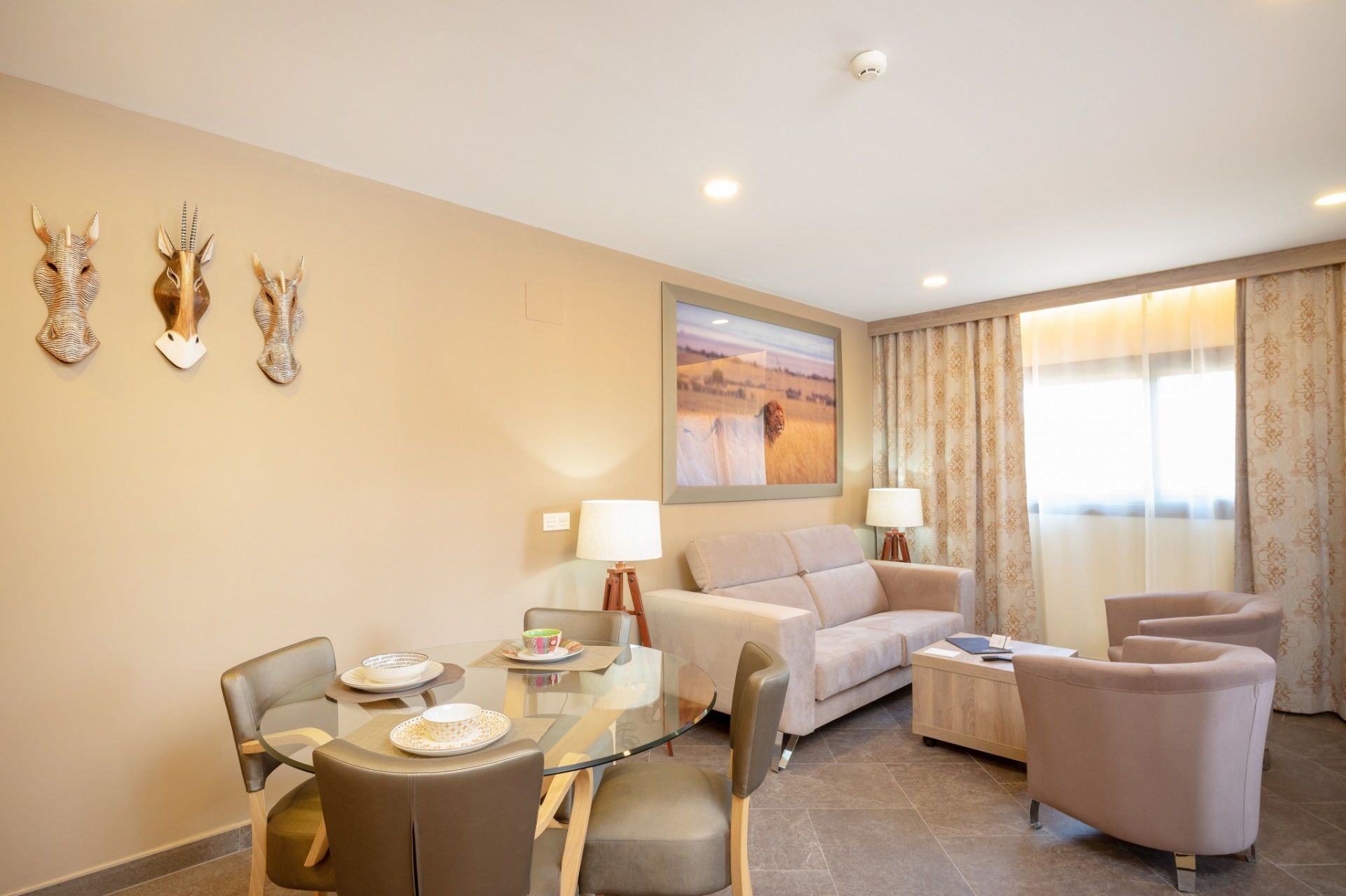 A modern hotel room with a glass dining table, a sofa, a chair, and a small side table.